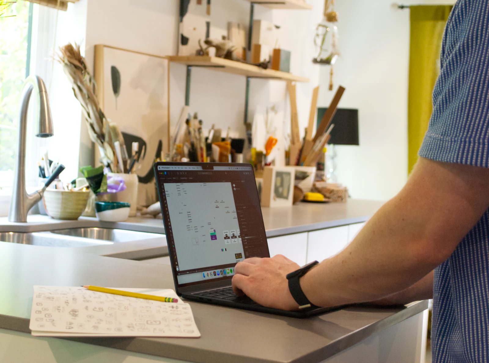 Ezrah working on a brand layout on a laptop at a kitchen counter, with sketches and a pencil beside it